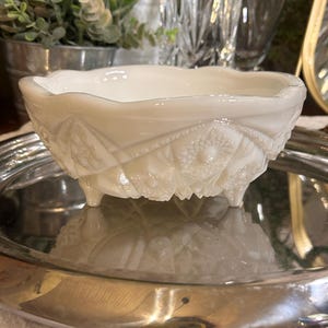May include: A white, vintage milk glass bowl with a scalloped rim and ornate embossed floral and geometric patterns. The bowl rests on three small feet and is displayed on a silver tray. A decorative piece for serving or display.