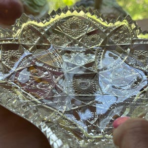 May include: A clear, cut-glass bowl with a detailed geometric pattern. The bowl has a scalloped edge and a diamond-like design throughout. The glass reflects light, creating a sparkling effect. The bowl is likely vintage and suitable for serving or display.