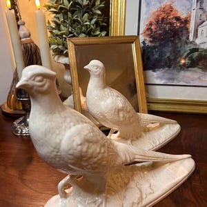 May include: A pair of off-white ceramic bird figurines, each depicted in a standing position on an integrated oval base. The birds feature detailed molded plumage, pointed beaks, and smooth, glossy surfaces, capturing a realistic avian form.