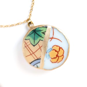 May include: A round pendant necklace featuring a ceramic shard with a gold-toned metal edge. The pendant displays a split design with a green leaf pattern on one side and a yellow floral motif on a white background on the other side.