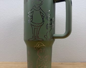 Puede incluir: Un vaso térmico verde con tapa y asa blancas. El vaso presenta imágenes grabadas de un personaje de dibujos animados con gorro de Papá Noel y su perro, junto con copos de nieve y otros diseños temáticos navideños. El vaso mide unos 25 cm de alto.