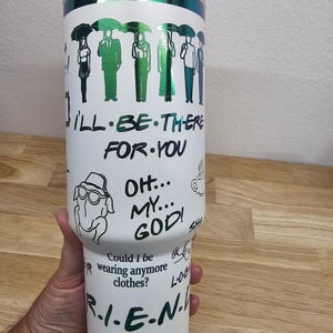 May include: White tumbler with a green metallic lid. The tumbler has a Friends theme with images of the characters, quotes, and the iconic New York City skyline. The text on the tumbler reads "I'll be there for you", "Oh my god", "Could I be wearing anymore clothes?" and "R.I.E.N."