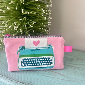 May include: Pink zippered pouch with a teal typewriter graphic and a pink heart. The pouch has a pink zipper and a pink fabric loop. The background is a light blue surface and a green and white decorative tree.