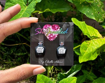May include: A pair of black poison bottle earrings with a skull and crossbones design. The earrings are on a white card with the text "She Jeweled" and "By: Chelle Alexis" and the social media handle "@ChelleAlexisArt".