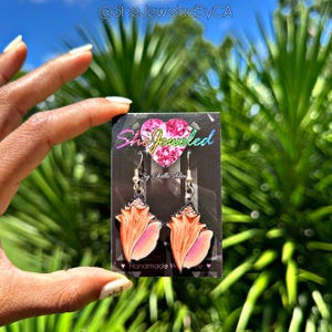 May include: A pair of seashell earrings with silver hooks, displayed on a black card. The earrings are orange and pink, with a detailed conch shell design. The card has the text "SheJeweled" and "By Chelle Alex".