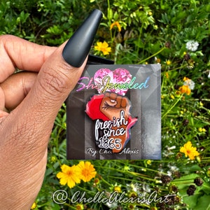 May include: A red, white, and blue enamel pin with a raised fist and the text "freeish since 1865" on a black background. The pin is on a card with the text "She Jeweled" and "By: Chel Alexis" on it.