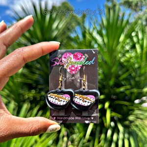 May include: Black heart-shaped earrings with a gold-colored gun design and white teeth on a pink background. The earrings are on a black card with the text "She Jeweled" and "Handmade With Love".