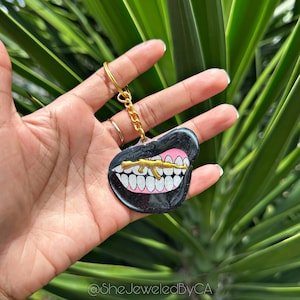 May include: A black and pink resin keychain shaped like a mouth with a gold-colored gun in it. The keychain is attached to a gold chain.