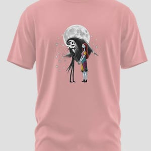 May include: A light pink t-shirt featuring a graphic of Jack Skellington and Sally from The Nightmare Before Christmas, holding hands in front of a full moon. The design includes stars and a dark silhouette of the characters.
