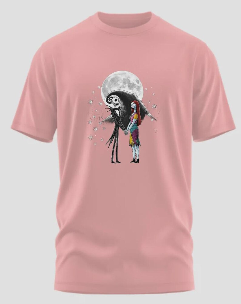 May include: A light pink t-shirt featuring a graphic of Jack Skellington and Sally from The Nightmare Before Christmas, holding hands in front of a full moon. The design includes stars and a dark silhouette of the characters.
