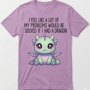 May include: A lilac-colored t-shirt featuring a cute cartoon dragon with large eyes and wings. The text on the shirt reads, "I FEEL LIKE A LOT OF MY PROBLEMS WOULD BE SOLVED IF I HAD A DRAGON."