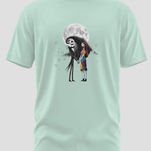 May include: A light green t-shirt featuring a graphic of Jack Skellington and Sally from The Nightmare Before Christmas, holding hands in front of a full moon. The design is in black and white with some color.