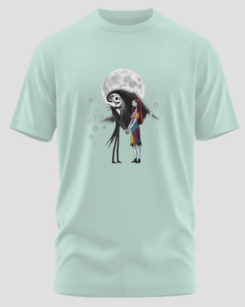 May include: A light green t-shirt featuring a graphic of Jack Skellington and Sally from The Nightmare Before Christmas, holding hands in front of a full moon. The design is in black and white with some color.