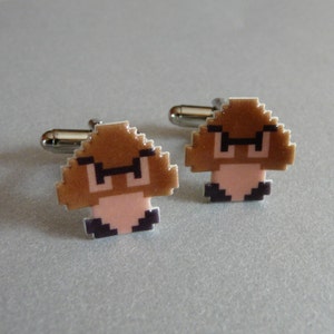 May include: A pair of pixelated Goomba cufflinks. The cufflinks feature a brown mushroom design with black-rimmed glasses and a pink face. The cufflinks have a silver-colored backing.