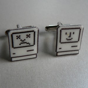 May include: A pair of cufflinks featuring a vintage Macintosh computer design. One cufflink has a happy face on the screen, while the other has a sad face.