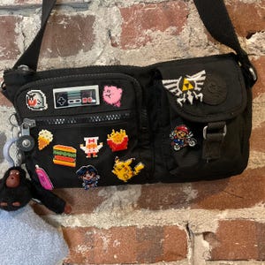 Könnte beinhalten: Eine schwarze Umhängetasche mit mehreren Taschen und einer Vielzahl von bunten gestickten Aufnähern, darunter ein Videospiel-Controller, eine Comicfigur und ein 'The Legend of Zelda'-Logo.