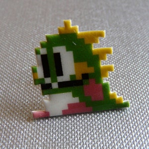 May include: A pixelated green and white creature pin with yellow accents. The pin features a retro video game character design with a pink tail and black eyes. The pin is a small, colorful accessory.