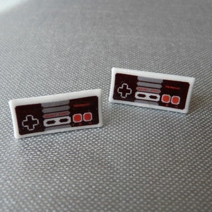 May include: A pair of rectangular earrings featuring a classic Nintendo controller design. The earrings have a white border with a dark brown controller layout, including a directional pad, buttons, and the Nintendo logo. The design is a nostalgic nod to retro gaming.
