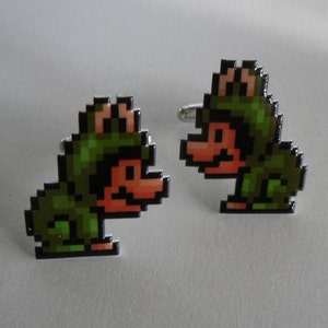 May include: A pair of pixelated cufflinks featuring a green and brown frog-like character. The cufflinks have a silver backing and a glossy finish. The design is reminiscent of retro video game graphics.