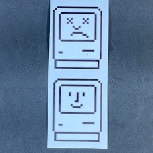 Puede incluir: Una pegatina blanca con dos diseños de ordenador pixelados. El ordenador superior tiene una cara triste con los ojos tachados, mientras que el ordenador inferior tiene una cara sonriente. La pegatina es rectangular y tiene una estética retro.