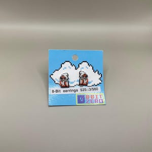 Puede incluir: Un par de pendientes pixelados de 8 bits en una tarjeta azul con un diseño de nube. Los pendientes presentan un diseño de personaje rojo y blanco. La tarjeta también incluye el texto "8-Bit earrings" y el logotipo "7 Bit Zero".