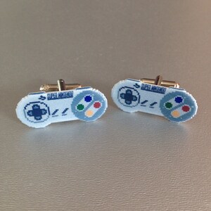 May include: A pair of cufflinks featuring a pixelated design of a Super Nintendo controller. The controllers are white with blue, green, yellow, and red buttons. The text "SUPER NINTENDO" is printed on each cufflink.