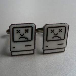 May include: A pair of novelty cufflinks featuring a pixelated computer design with a sad face. The cufflinks are white with black detailing and silver-toned metal hardware. A unique accessory for computer enthusiasts.