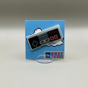 May include: A retro-style pin featuring a pixelated design of a classic video game controller. The controller is black, gray, and red, set against a blue background with a white cloud. The pin has the text "8-BIT ZERO".