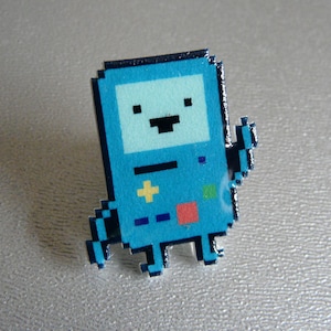 May include: A pixelated, light blue pin featuring a character with a square head, black eyes, and a black mouth. The character has a yellow plus sign and a red square on its body. The pin is a fun accessory.