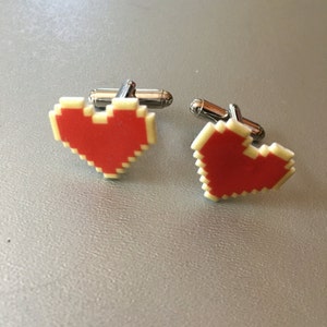 May include: A pair of silver cufflinks with red pixelated heart designs. The hearts are outlined in white.
