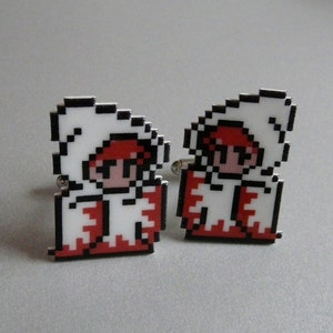 Può includere: Un paio di gemelli con un design pixelato di un personaggio che indossa una tunica bianca con cappuccio e abiti rossi.