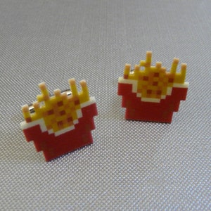May include: A pair of pixelated French fry earrings. Each earring features a red carton filled with yellow fries, with a white border. The earrings have a retro video game aesthetic.