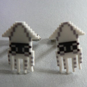 May include: A pair of white cufflinks shaped like pixelated space invaders with black eyes. The cufflinks are made of a white plastic material and have a silver metal backing.
