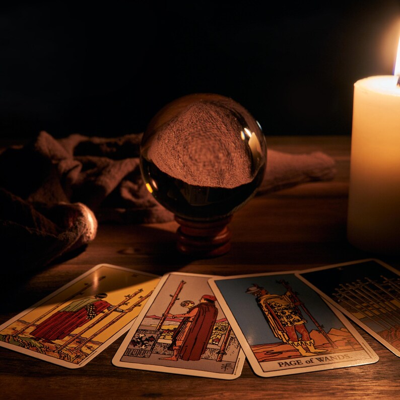 May include: Four tarot cards with illustrations of people holding staffs, laid out on a wooden table in front of a crystal ball and a lit candle. The cards are from the "Wands" suit and include the Page of Wands.