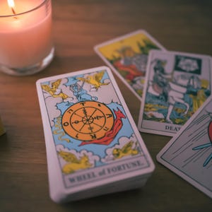 May include: A tarot card reading with a lit candle in the background. The card in the foreground is the Wheel of Fortune card, which features a wheel with a red and yellow design and a snake. The card is surrounded by other tarot cards, including Death and The Chariot.