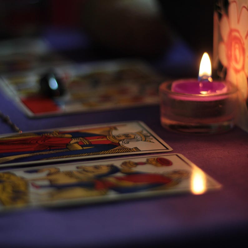 May include: Tarot cards laid out on a purple surface with a lit candle in a glass holder. The cards feature illustrations of people and symbols.