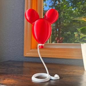 May include: A bright red, three-dimensional balloon sculpture shaped like a mouse head with two rounded ears. The balloon is attached to a white, coiled rope-like base that extends upward to connect to the bottom of the red balloon structure.
