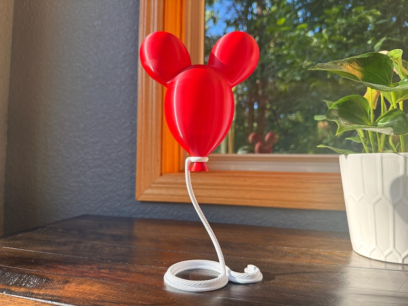 May include: A bright red, three-dimensional balloon sculpture shaped like a mouse head with two rounded ears. The balloon is attached to a white, coiled rope-like base that extends upward to connect to the bottom of the red balloon structure.