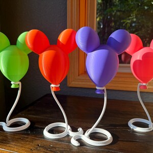 May include: Four decorative balloon-shaped ornaments with rounded ears, each featuring a distinct solid color: lime green, orange, purple, and pink. Each balloon is attached to a white, curved, rope-textured stand with a circular base.
