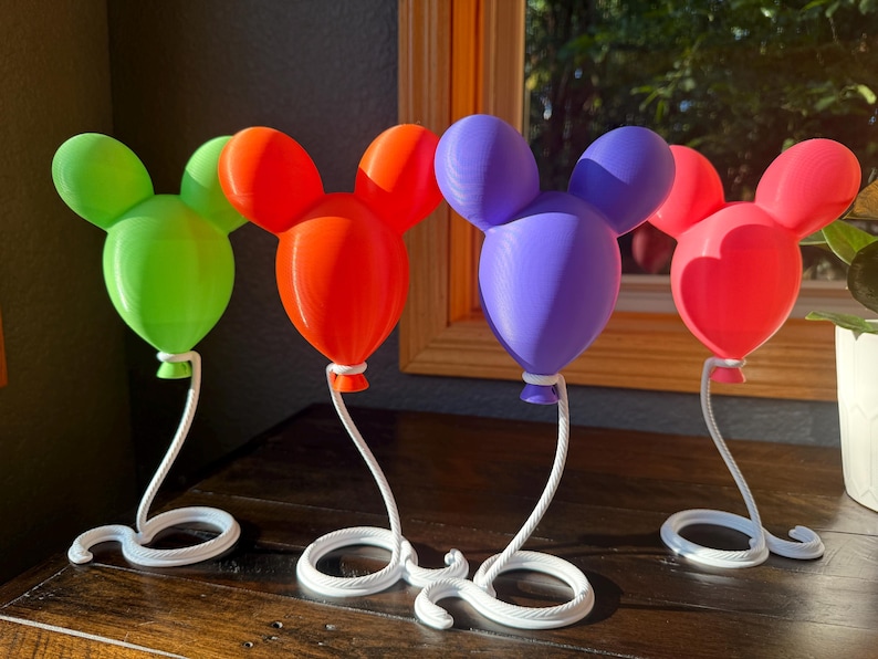 May include: Four decorative balloon-shaped ornaments with rounded ears, each featuring a distinct solid color: lime green, orange, purple, and pink. Each balloon is attached to a white, curved, rope-textured stand with a circular base.