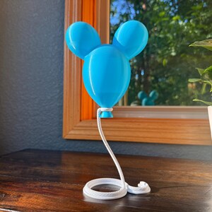May include: A bright blue balloon-shaped sculpture with two rounded ears at the top. The balloon is attached to a white, coiled rope-like base that extends upward to support the balloon, creating a continuous, connected structure.