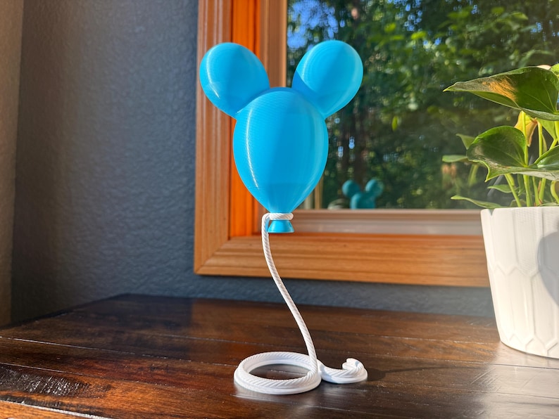 May include: A bright blue balloon-shaped sculpture with two rounded ears at the top. The balloon is attached to a white, coiled rope-like base that extends upward to support the balloon, creating a continuous, connected structure.