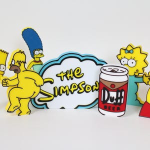 May include: A set of yellow cartoon characters from The Simpsons, including Bart, Homer, Marge, Lisa, and Maggie. The characters are standing in front of a white sign that says "The Simpsons" and a red can of Duff beer.