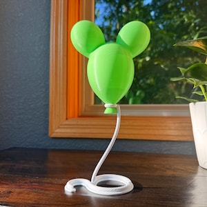 May include: A bright lime green balloon-shaped ornament with two rounded ears at the top, attached to a white, coiled, rope-textured base that extends upward as a thin, curved stem to support the balloon from its bottom knot.