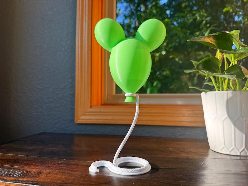 May include: A bright lime green balloon-shaped ornament with two rounded ears at the top, attached to a white, coiled, rope-textured base that extends upward as a thin, curved stem to support the balloon from its bottom knot.