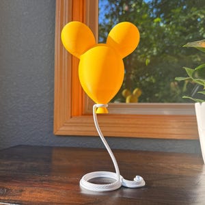 May include: A bright yellow, three-dimensional balloon sculpture shaped like a mouse head with two rounded ears. The balloon is attached to a white, coiled rope base that extends upward to connect to the bottom of the yellow balloon structure.