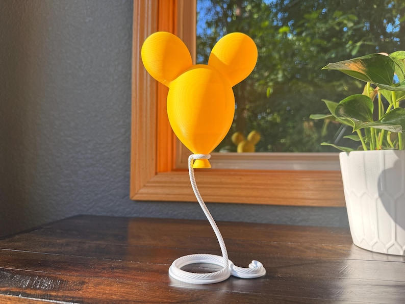 May include: A bright yellow, three-dimensional balloon sculpture shaped like a mouse head with two rounded ears. The balloon is attached to a white, coiled rope base that extends upward to connect to the bottom of the yellow balloon structure.