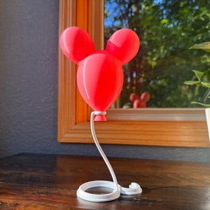 May include: A bright red, three-dimensional balloon sculpture shaped like a mouse head with two rounded ears. The balloon is attached to a white, coiled rope base that extends upward to support the balloon, creating a floating appearance.