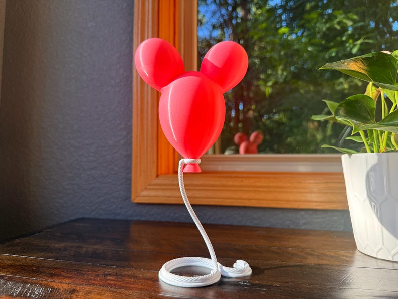 May include: A bright red, three-dimensional balloon sculpture shaped like a mouse head with two rounded ears. The balloon is attached to a white, coiled rope base that extends upward to support the balloon, creating a floating appearance.