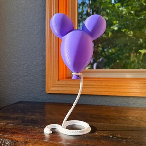 May include: A purple 3D-printed balloon sculpture featuring a rounded head with two circular ears, attached to a white, coiled, rope-like base that extends upward to support the balloon from its bottom knot, creating a floating effect.